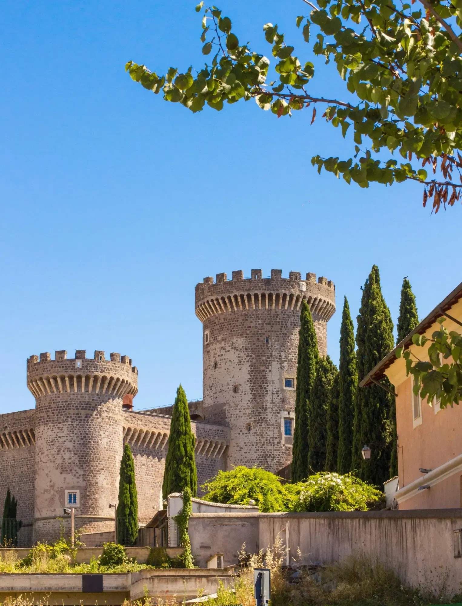 Zwei steinerne Türme der Burg Rocca Pia in Tivoli vor einem klaren blauen Himmel, mit Zypressenbäumen in der Nähe.