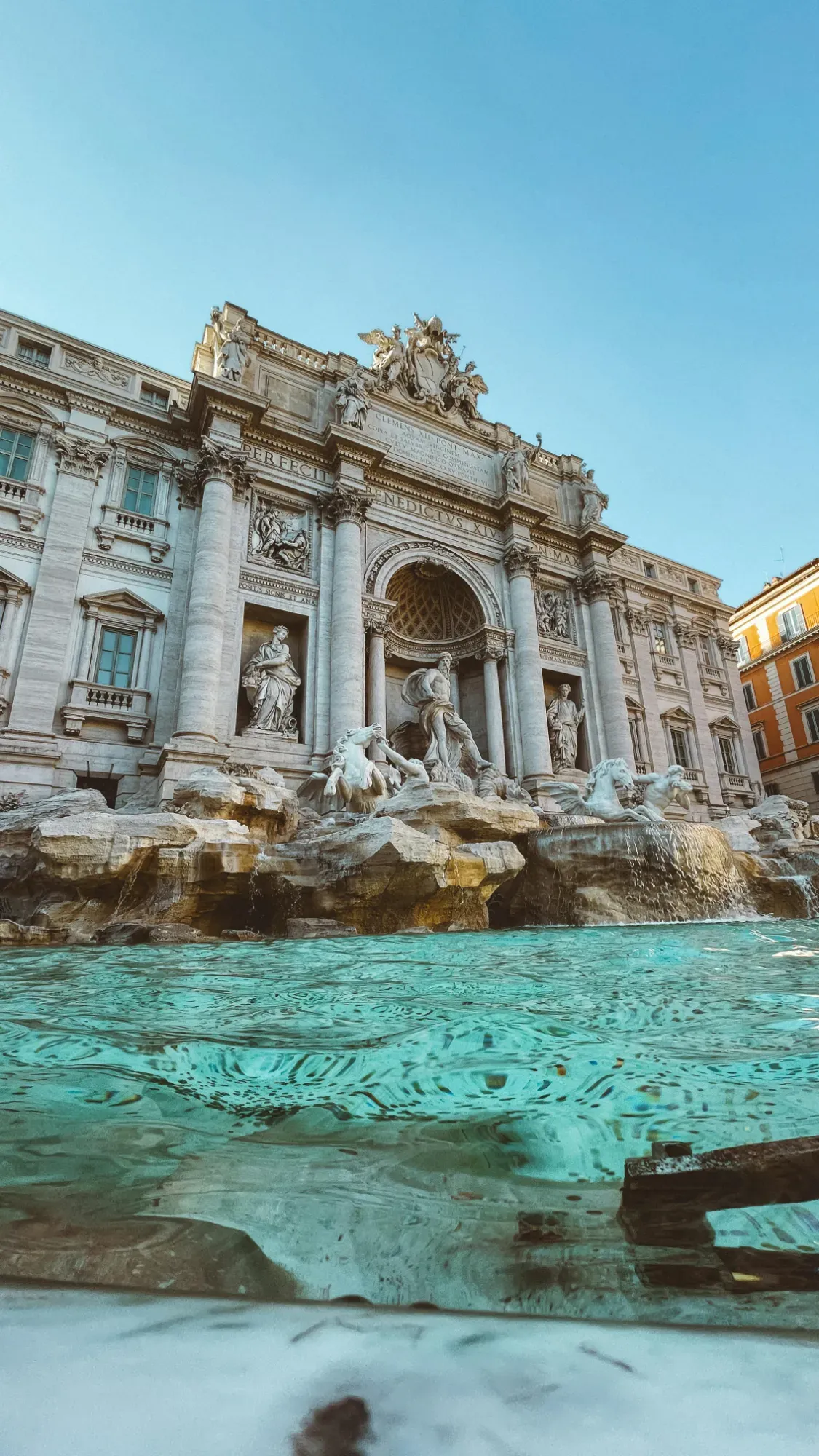 A wide shot of the Trevi Fountain in Rome, showing its ornate facade, sculptures, and clear blue water with reflections.