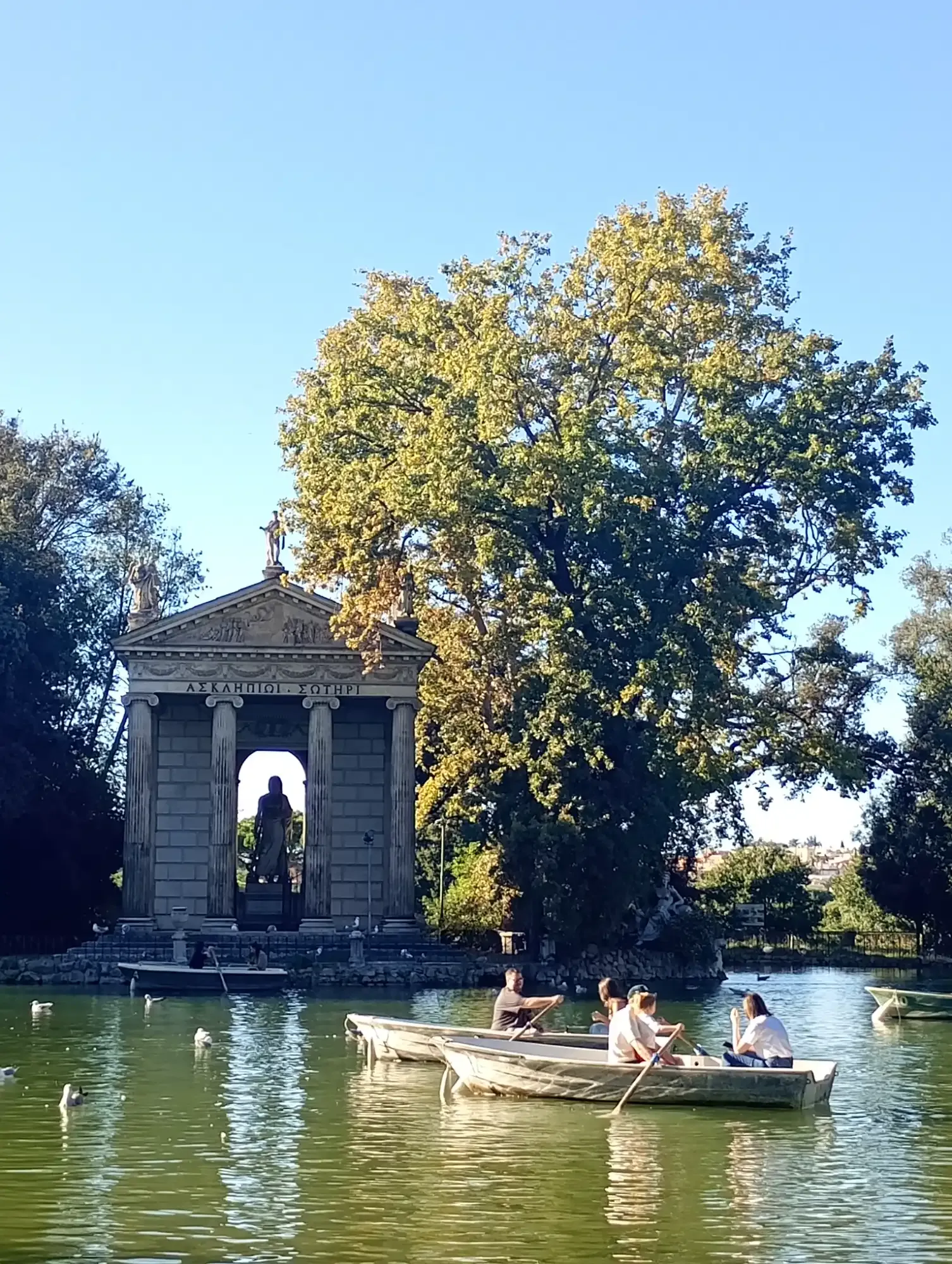 Classical temple with columns and statues by a pond with people rowing boats in Rome's Villa Borghese.
