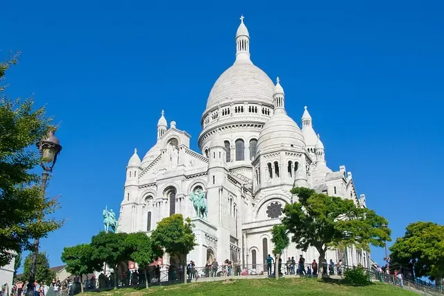 Die weiße Basilika Sacré-Cœur in Paris vor klarem blauem Himmel, mit Menschen auf einem grasbewachsenen Hügel davor.