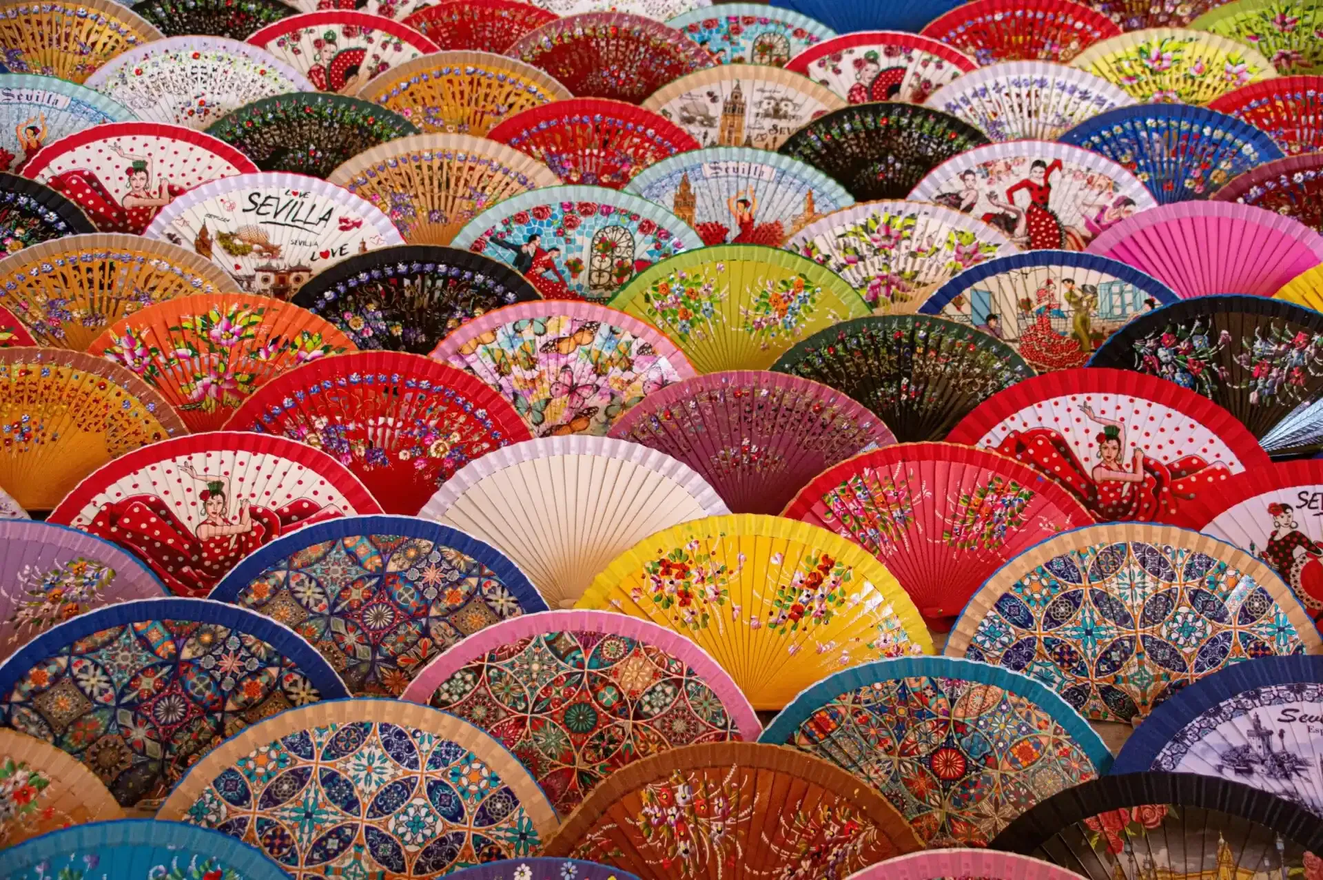 Close-up of numerous colorful Spanish hand fans arranged in rows, featuring various patterns like flamenco dancers, flowers,
