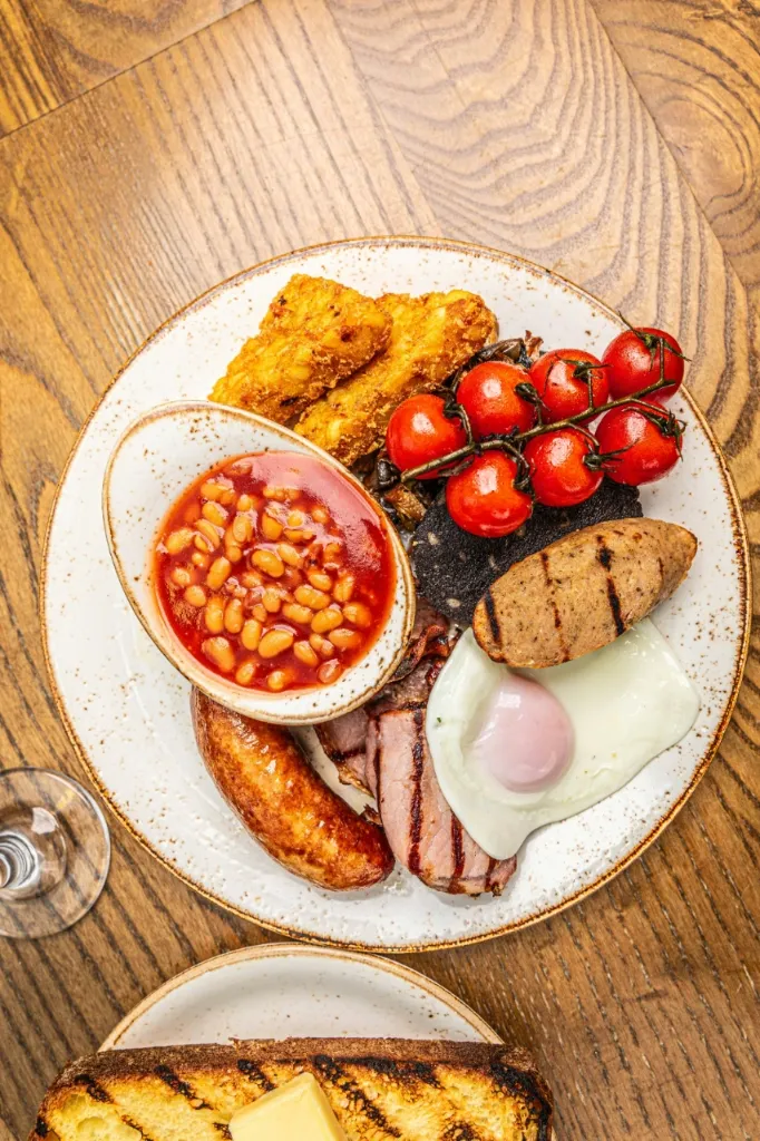 Desayuno inglés completo en un plato blanco con frijoles horneados, hash browns, tomates cherry, morcilla, salchicha, tocino