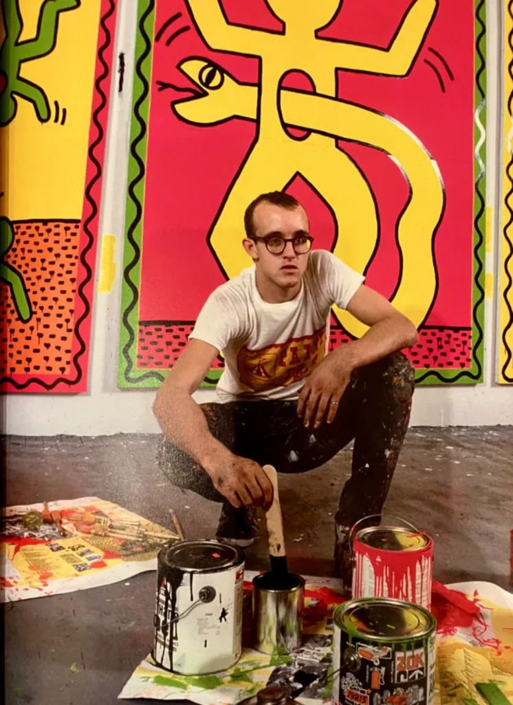 Keith Haring crouches in a studio surrounded by his colorful pop art paintings, paint cans, and brushes.