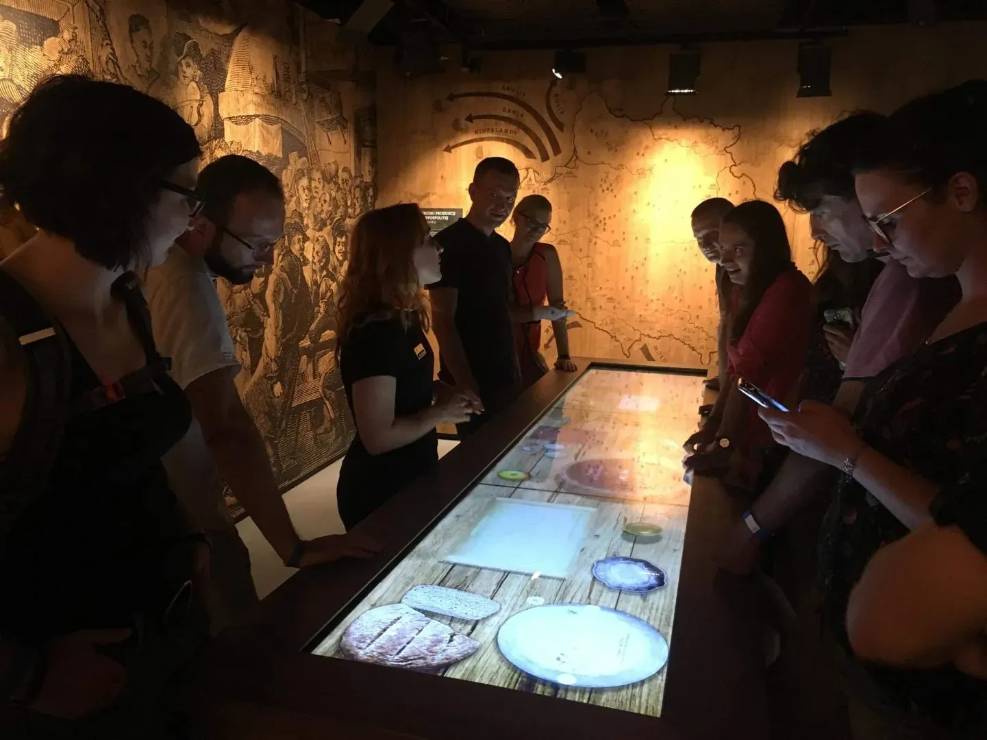 Group of people looking at an illuminated interactive display table with food graphics in a dimly lit museum room.