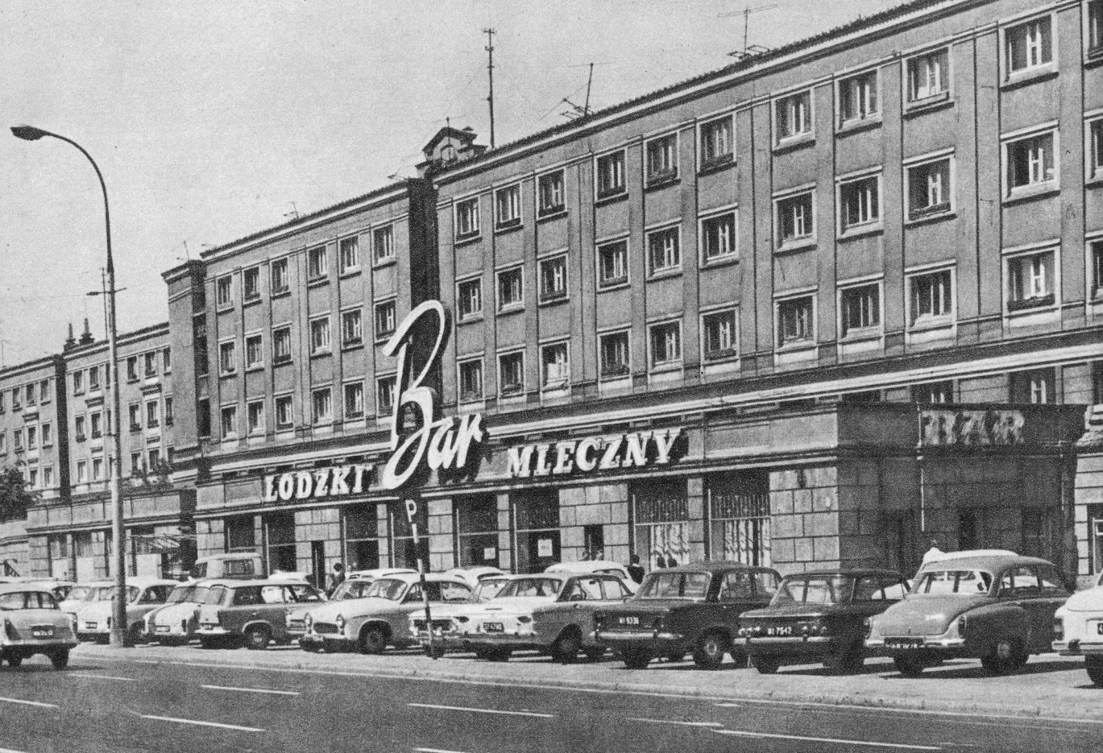 Ulica w Warszawie z budynkiem Bar Mleczny i jego dużym szyldem. Wzdłuż krawężnika zaparkowanych jest kilka klasycznych samoch