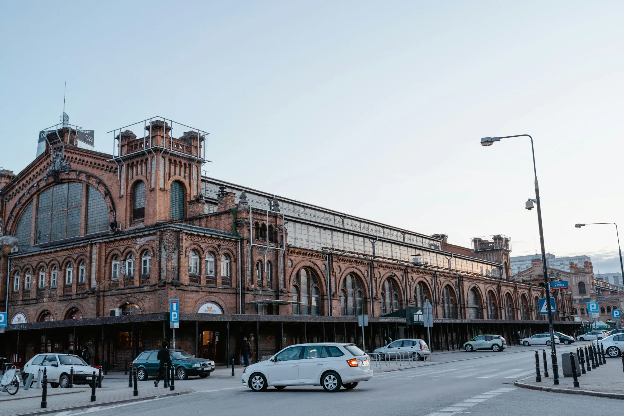 Large red brick market hall building in Warsaw with arched windows and a glass roof. Cars are parked on the street in front.