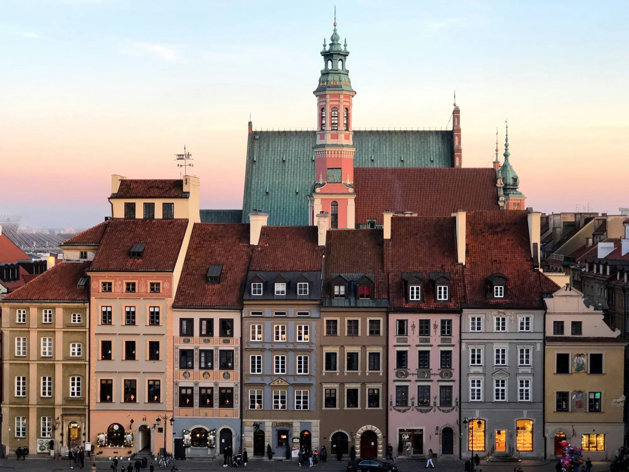 Bunte historische Bürgerhäuser in der Warschauer Altstadt bei Sonnenuntergang, mit Kirchturm im Hintergrund.