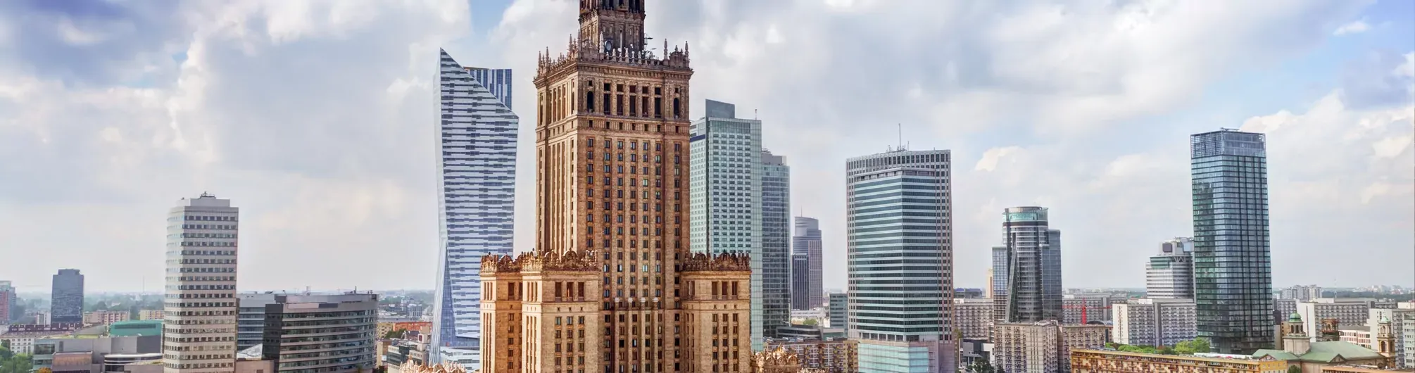 View of the Palace of Culture and Science, a tall brown building, surrounded by modern glass skyscrapers under a cloudy sky.