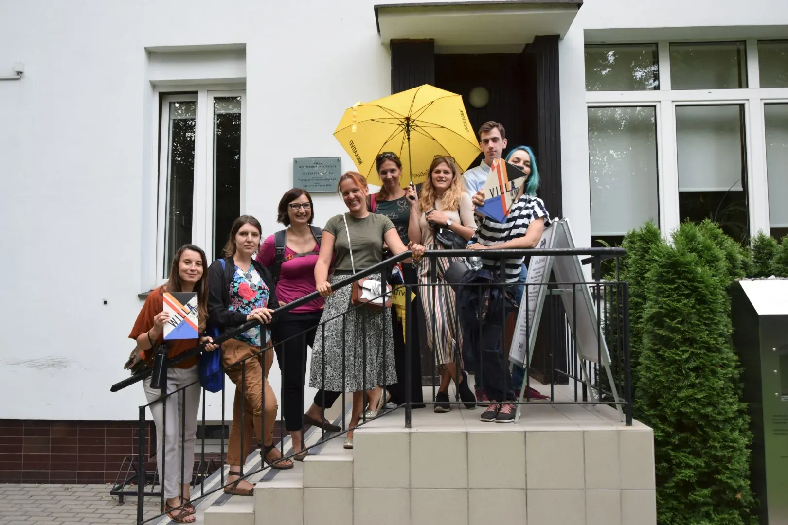 A group of people standing on the outdoor steps of a white building. Some hold books; a yellow umbrella is open overhead.