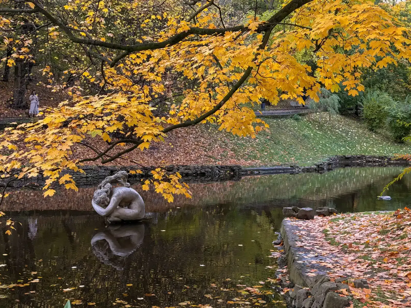 Escultura de piedra de una mujer en el estanque de un parque bajo ramas otoñales de arce.