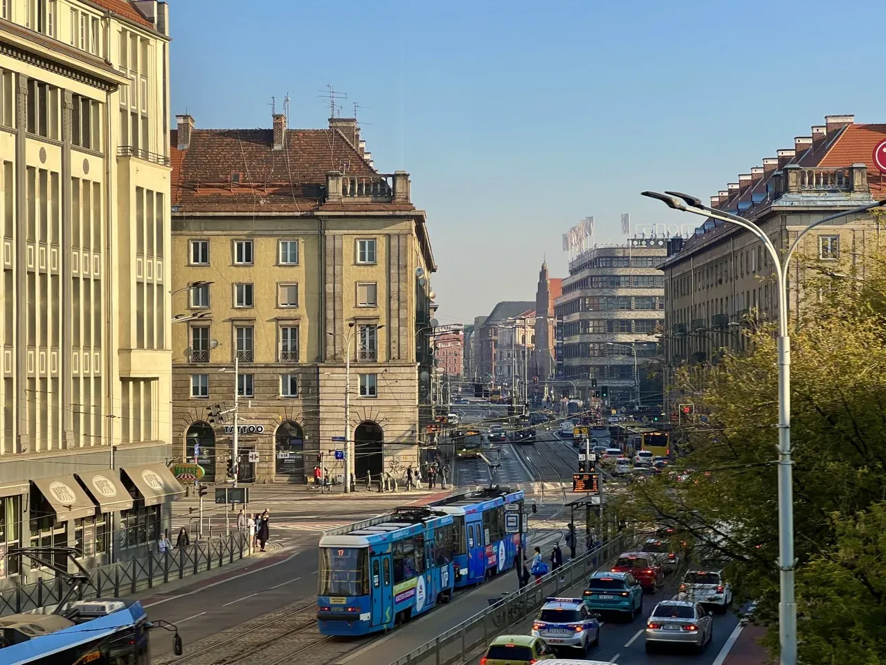 A wide city street with trams, cars, and pedestrians, flanked by multi-story buildings under a clear sky.