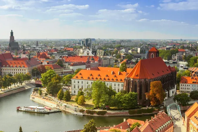 Aerial view of Wroclaw cityscape with Odra River, historic buildings, bridges, and Wrocław Cathedral under a blue sky.