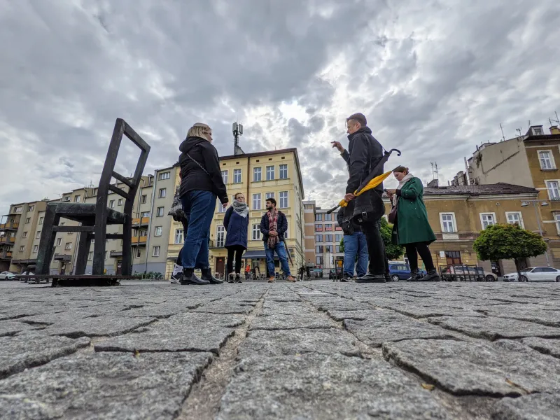 Grupa turystów na wycieczce z przewodnikiem w Krakowie, zwiedzająca plac miejski z dużą brązową rzeźbą krzesła.