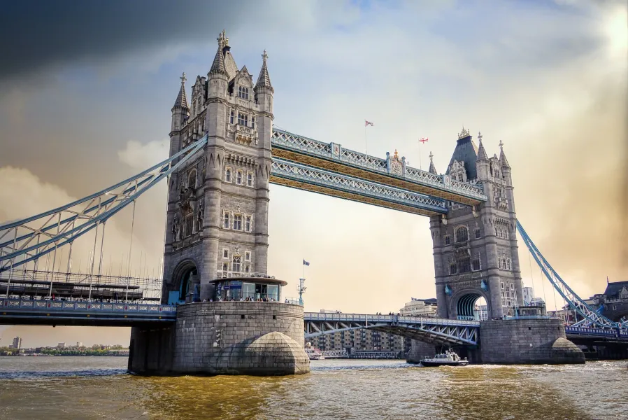 Most Tower Bridge rozciągający się nad szeroką rzeką pod częściowo zachmurzonym niebem, z łodzią na wodzie i budynkami w tle.