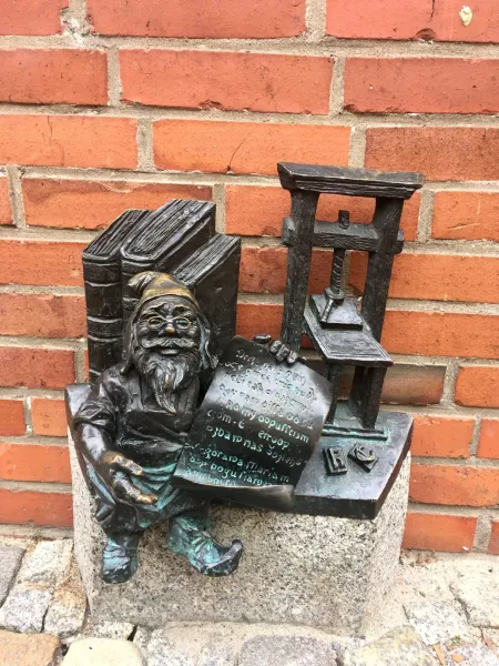 Brązowa figurka krasnala we Wrocławiu, siedząca przy prasie drukarskiej i książkach.