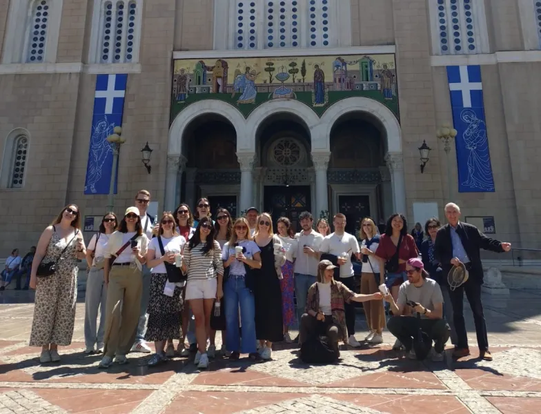 A tour group smiles for a photo in front of the Metropolitan Cathedral of Athens on a sunny day.