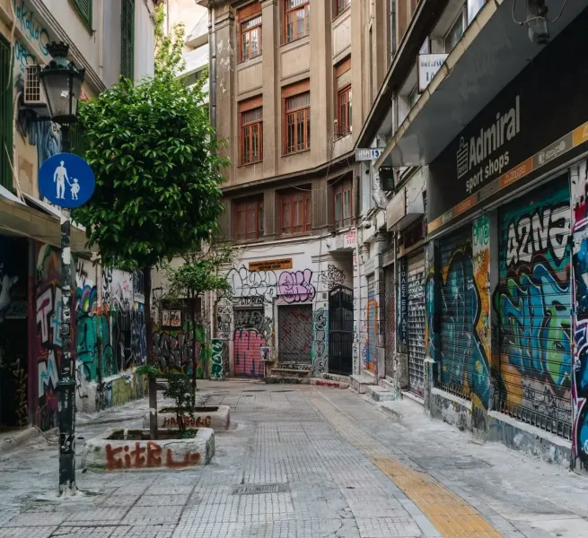 Narrow urban alley with buildings and shop shutters covered in colorful graffiti. Small trees in planters and a pedestrian...