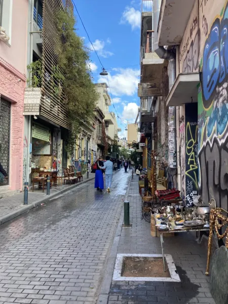 Paved street in Athens with buildings covered in graffiti and a flea market stall on the right. Two people walk away.