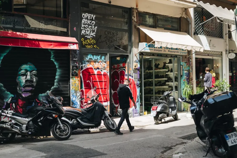 A sunny street in Athens with a large mural on a shop shutter, parked motorcycles, and a person walking past storefronts.