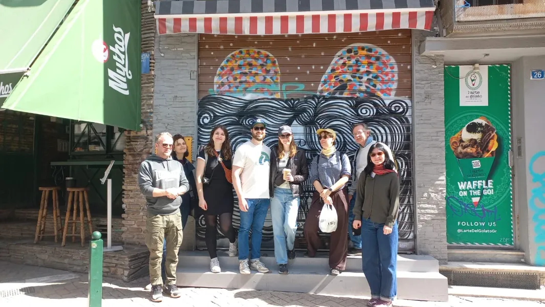 A group of tourists poses for a photo in front of a closed shop with graffiti on the streets of Athens.