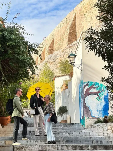 Three people on stone stairs in Athens, one with a yellow umbrella. A building with a tree mural and a large stone wall ar...