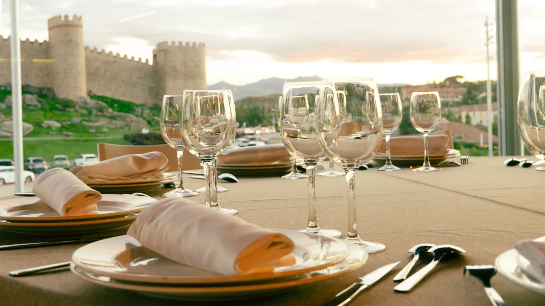 Una mesa de comedor con copas y platos, con vistas a las murallas medievales de Ávila, España, al atardecer.
