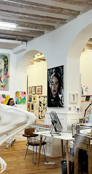 Wnętrze pracowni artystycznej z obrazami na białych ścianach, drewnianymi belkami, białą rzeźbą i kobietą obserwującą sztukę.