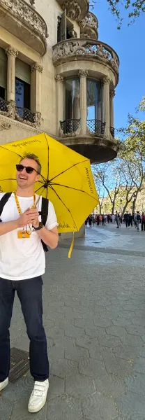 Guía turístico sonriente con paraguas amarillo de Walkative en una calle soleada de Barcelona. Edificio modernista ornamen...