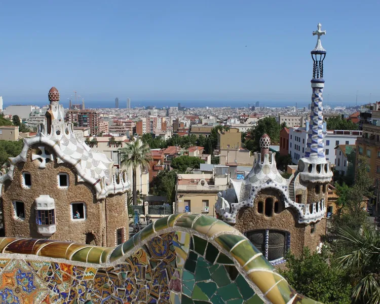 Blick vom Park Güell auf zwei Gaudi-Gebäude mit Mosaikdächern, eine bunte Fliesenmauer und Barcelonas Stadtbild mit Meer.