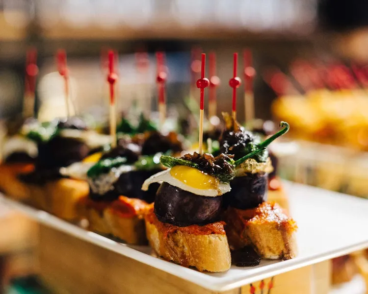 Nahaufnahme eines Tellers Pintxos, kleiner offener Sandwiches mit verschiedenen Belägen, gehalten von roten Zahnstochern. ...