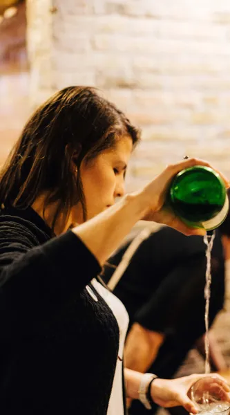 Persona sirviendo sidra de una botella verde en un vaso, creando espuma. Pared de piedra borrosa al fondo.
