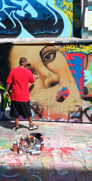 Osoba w czerwonej koszulce malująca sprayem duży mural twarzy kobiety na ścianie pokrytej graffiti. Puszki z farbą na ziemi.