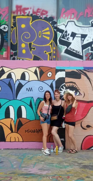 Trzy uśmiechnięte kobiety stojące przed kolorową ścianą pokrytą abstrakcyjnym graffiti i dużym muralem ze stylizowanymi twarz