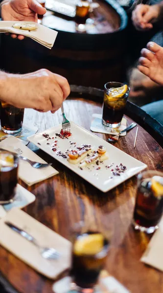 Hände von Personen an einem Holztisch mit Tapas und Getränken. Eine Person isst ein Gericht mit einer Gabel.