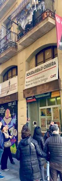 Grupa ludzi na wycieczce ulicznej patrzy w górę na budynek ze znakami "Solidaridad Obrera" i "CNT" oraz graffiti na roletach.