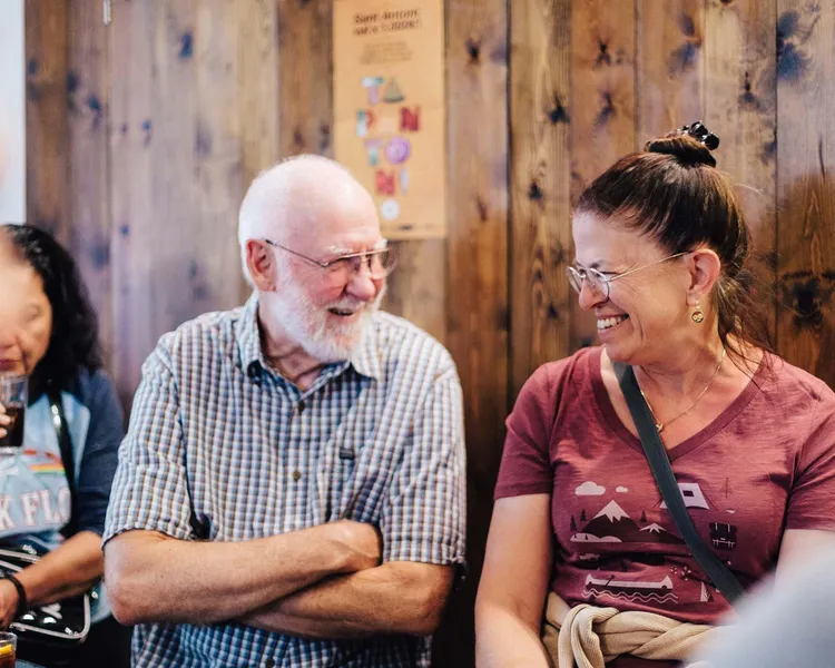 Dos viajeros sonrientes, un hombre mayor y una mujer, conversando en un ambiente informal de bar con pared de madera y cartel