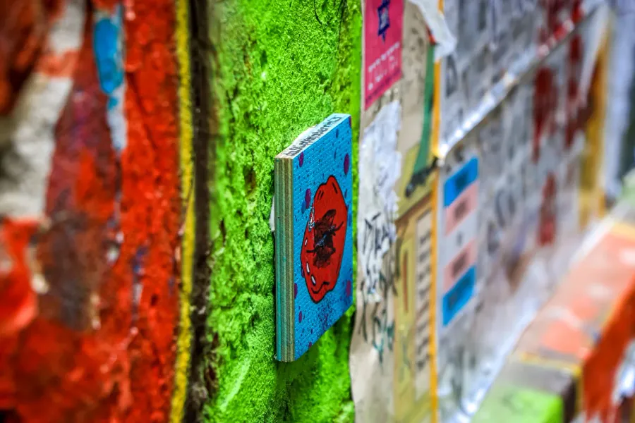 Close-up of a small wooden artwork on a bright green textured wall, surrounded by layers of paint and peeling posters.