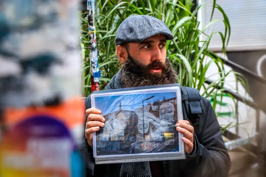 A tour guide with a beard and flat cap holds up a photograph of a large street art mural in Berlin.