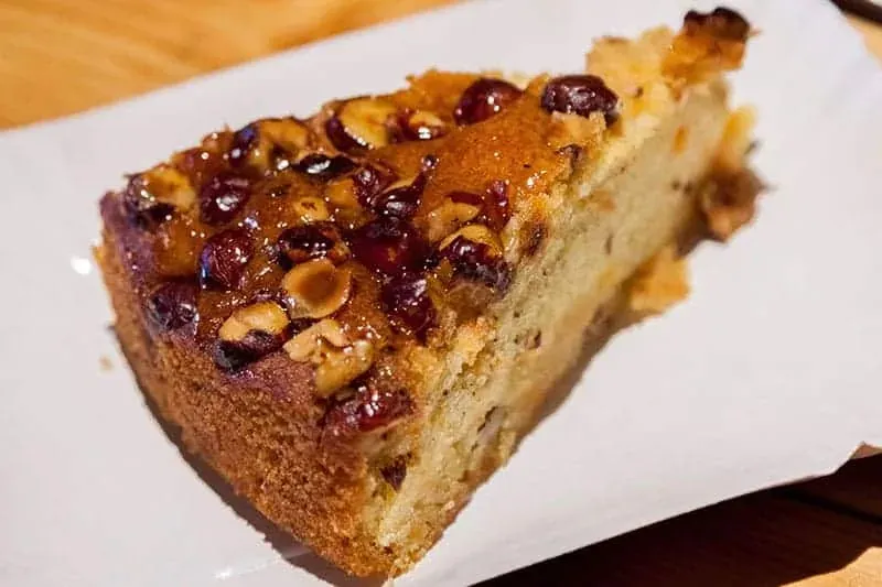 Ein Stück Kuchen mit glänzender Glasur, gerösteten Haselnüssen und getrockneten Beeren auf einem weißen Teller.