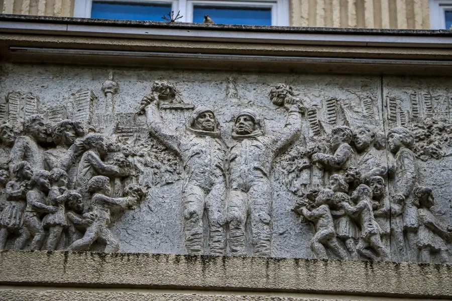 Detailed stone relief sculpture depicting two cosmonauts holding bouquets, surrounded by adults and children, with the Ber...