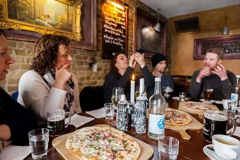 Eine Gruppe von Leuten sitzt an einem langen Tisch in einem gemütlichen Berliner Restaurant, mit Flammkuchen und Getränken.