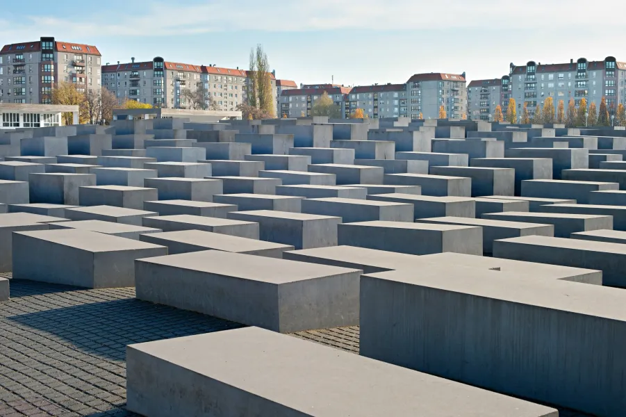Field of concrete stelae of varying heights, forming paths, with modern apartment buildings and trees in the background.