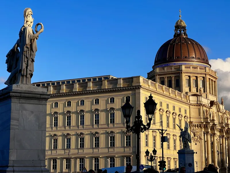 Estatua clásica de piedra de una mujer con corona, con la cúpula y fachada del Foro Humboldt en Berlín detrás.