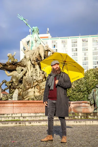 Hombre barbudo con gorra y paraguas amarillo de Walkative, de pie frente a la Fuente de Neptuno en Berlín.