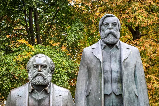 Bronze statues of Karl Marx and Friedrich Engels in Berlin, with green and autumnal trees in the background.