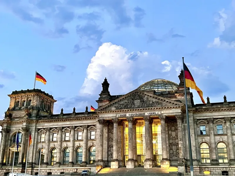 The Reichstag Building in Berlin at dusk, with German flags waving under a cloudy blue and white sky.