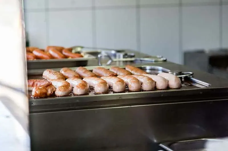 Würstchen braten auf einer Metallplatte in einer Küche mit weißen Fliesen. Eine Grillzange ist sichtbar.