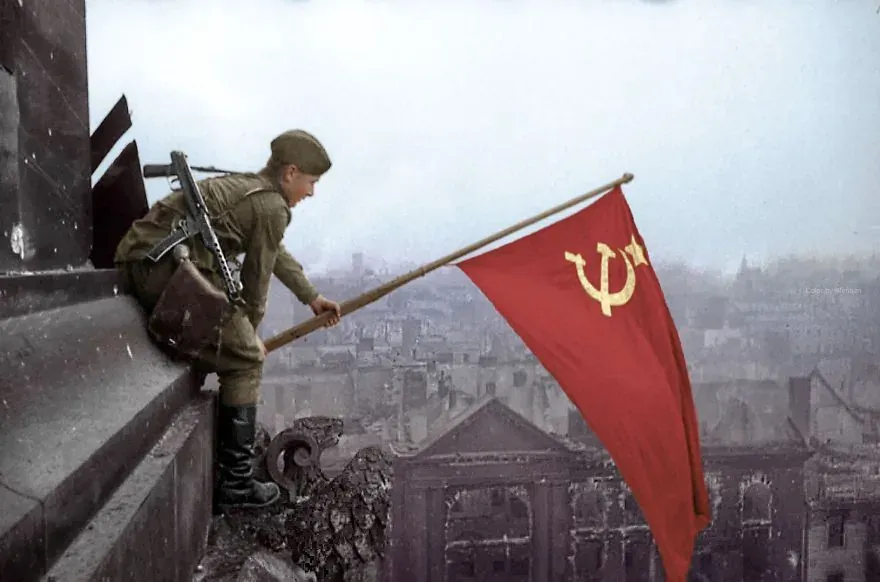 Soviet soldier planting a red flag with hammer and sickle on a destroyed building, city ruins in background.