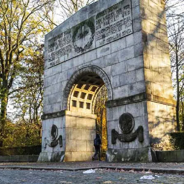 Large grey stone archway monument with carved inscriptions and reliefs, a person walks through it in a park setting.