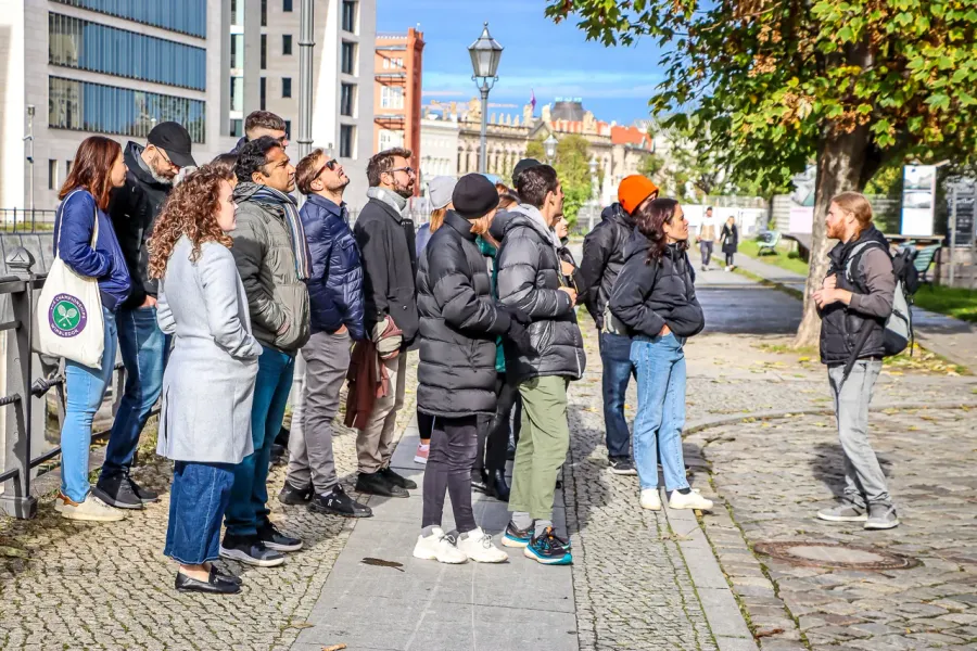 A group of tourists stands on a cobbled street listening to a tour guide, with modern and historic buildings in the backgr...
